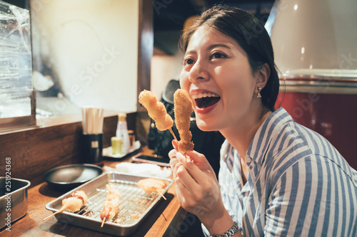 young girl cheerfully trying japanese dish of deep-fried skewered meat and vegetables in izakaya. woman smiling eating kushikatsu in pub sitting at counter at night after long day travel have dinner photo