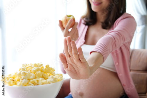 Dieting pregnancy woman refusing her favorite junk food such as popcorn by raising her hand and pushing it and choosing green apple instead for having a good health for her newborn baby in the future. photo