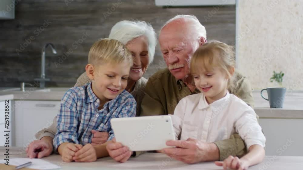 Lockdown of aged couple sitting at kitchen table with little girl and boy, holding tablet, watching pictures in internet and discussing them Lockdown of aged couple sitting at kitchen table with little girl and boy, holding tablet, watching pictures in internet and discussing them