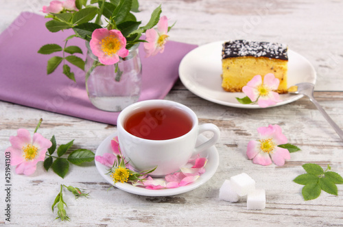 Cup of tea with cheesecake and wild rose flower on rustic board