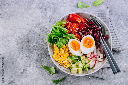 Healthy vegan lunch bowl. Avocado, egg, red bean, tomato, radish, corn, green leaves vegetables salad. Concrete background. Top view. Copy space