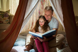 Family quality time. Father and daughter sit in homemade pink tent with flowers, read big book, look at each other, smile and laugh. Cozy stylish room. Family bonds concept 