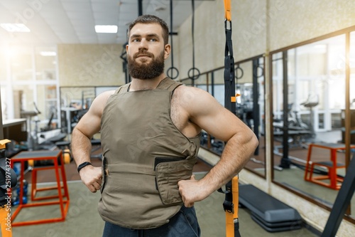 Portrait muscular caucasian bearded man dressed in weighted vest in the gym, military style