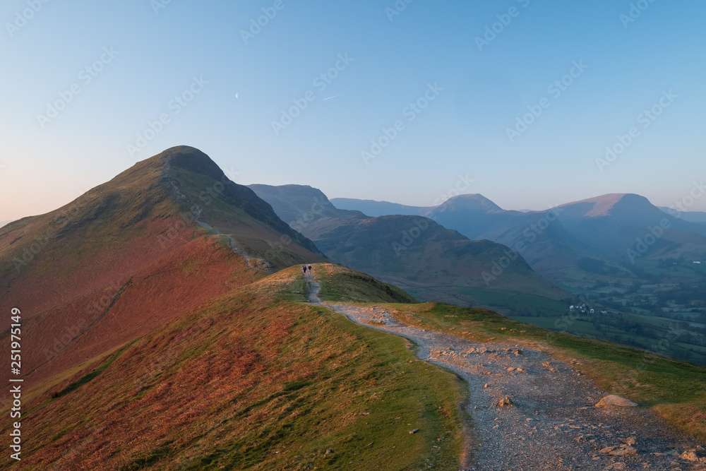 Two people in the distance walking down the footpath from Cat Bells as first light catches the fells,  Hindscarth and the Newlands Valley are in the background, Lake District, UK