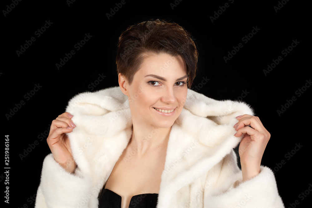 The young beautiful woman in a white fur coat on a black background looks into the camera. The young beautiful woman in a white fur coat on a black background looks into the camera.