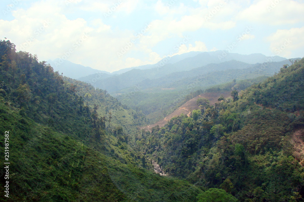 The landscape of numerous mountain ranges covered by green forest under the rays of the midday sun on the background of a cloudy blue sky on the horizon.