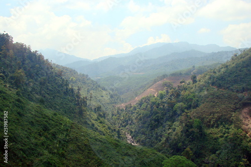 The landscape of numerous mountain ranges covered by green forest under the rays of the midday sun on the background of a cloudy blue sky on the horizon.