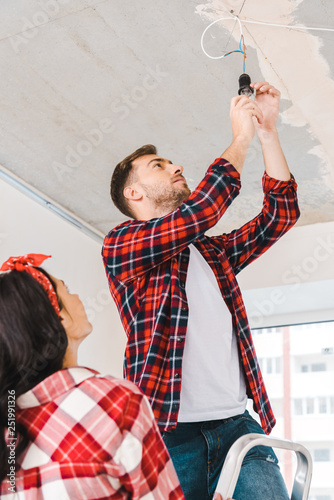 handsome man standing on ladder and chanding light bulb near girlfriend photo
