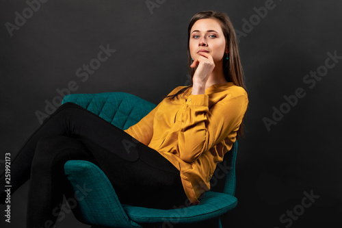 A young girl with brown hair sits on a turquoise chair in a yellow shirt and leggings, lifting her legs up. Sexy portrait in the studio © Julia