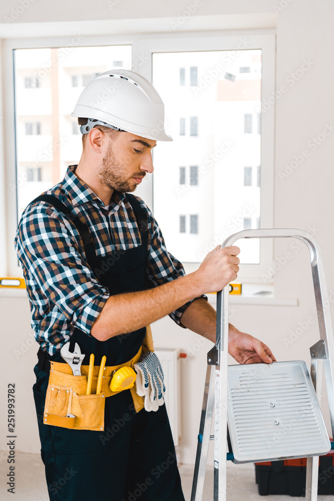 handsome handyman in helmet holding ladder in room handsome handyman in helmet holding ladder in room