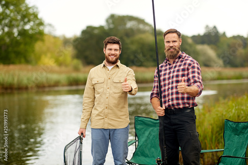 leisure and people concept - happy friends with fishing rods on lake showing thumbs up