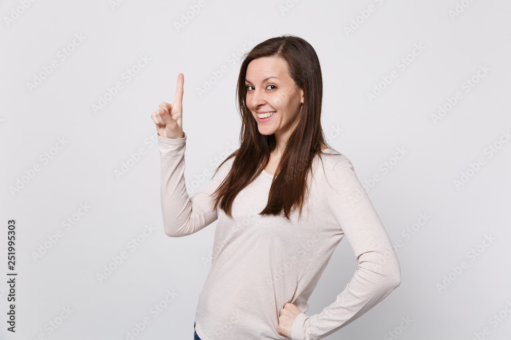 Portrait of smiling beautiful young woman in light clothes standing pointing index finger up isolated on white wall background in studio. People sincere emotions lifestyle concept. Mock up copy space. Portrait of smiling beautiful young woman in light clothes standing pointing index finger up isolated on white wall background in studio. People sincere emotions lifestyle concept. Mock up copy space.