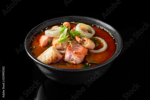 Hot miso soup with salmon, squids, scallops, seaweed, mushrooms, herbs in bowl on black background. Japanese food.