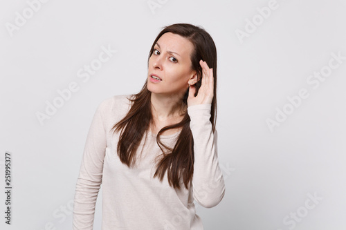 Portrait of concerned young woman in light clothes looking camera and eavesdrop with hearing gesture isolated on white wall background. People sincere emotions, lifestyle concept. Mock up copy space.