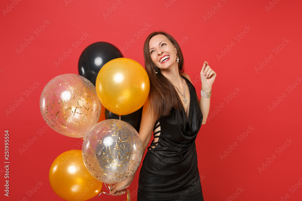 Naklejka premium Smiling young girl in little black dress celebrating looking up holding air balloons isolated on bright red background. International Women's Day, Happy New Year birthday mockup holiday party concept.