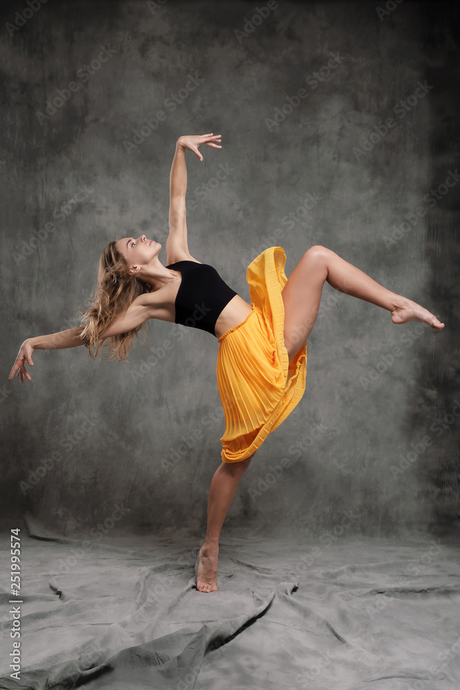 A beautiful graceful slender female dancer performs choreographic figures and movements on a gray dark fabric background.