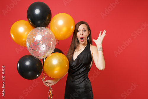 Shocked young girl in little black dress celebrating spreading hands, holding air balloons isolated on red background. International Women's Day, Happy New Year, birthday mockup holiday party concept. © ViDi Studio