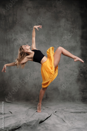 Fototapeta Naklejka Na Ścianę i Meble -  A beautiful graceful slender female dancer performs choreographic figures and movements on a gray dark fabric background.