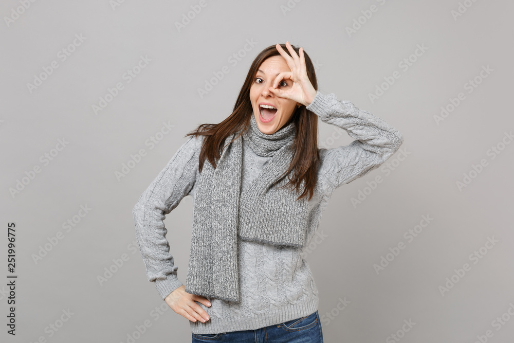 Excited young woman in gray sweater, scarf showing OK gesture near eyes isolated on grey wall background. Healthy fashion lifestyle, people sincere emotions, cold season concept. Mock up copy space. Excited young woman in gray sweater, scarf showing OK gesture near eyes isolated on grey wall background. Healthy fashion lifestyle, people sincere emotions, cold season concept. Mock up copy space.