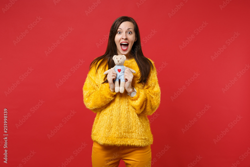 Excited young woman in yellow fur sweater keepin mouth wide open holding teddy bear plush toy isolated on red wall background in studio. People sincere emotions, lifestyle concept. Mock up copy space. Excited young woman in yellow fur sweater keepin mouth wide open holding teddy bear plush toy isolated on red wall background in studio. People sincere emotions, lifestyle concept. Mock up copy space.