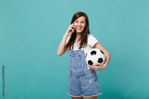 Beautiful happy young girl football fan with soccer ball talking on mobile phone, conducting pleasant conversation isolated on blue turquoise background. People emotions, sport family leisure concept. © ViDi Studio