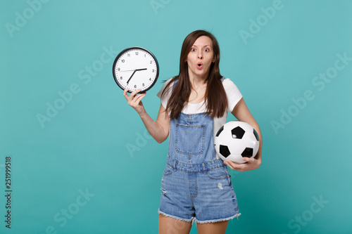 Shocked young woman football fan holding soccer ball, round clock isolated on blue turquoise wall background in studio. Time is running out. People emotions, sport family leisure lifestyle concept.