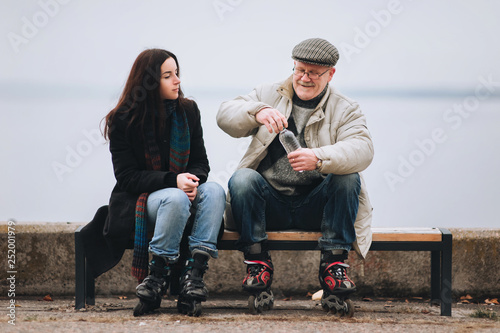 Father and daughter on roller skates sit on a bench. An elderly man has fun skating on roller skates. Happy pension. Active old people.