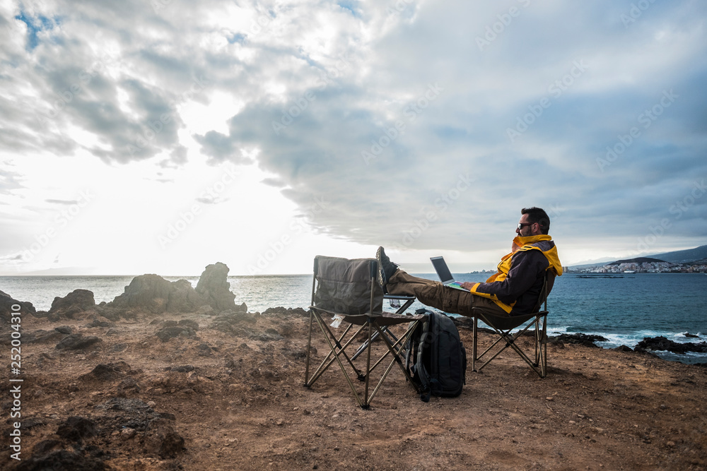 Digital nomad and traveler middle age adult man sitting and working with internet and laptop in totally fredom in front of the ocean - backpacker lifestyle and alternative office for happy life - obrazy, fototapety, plakaty 