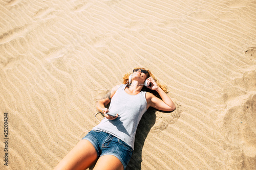 Pretty girl with curly hair and short jeans enjoying the sand and the beach listining music from phone and headphone viewed from above - concept of summer vacation and happy lifestyle photo