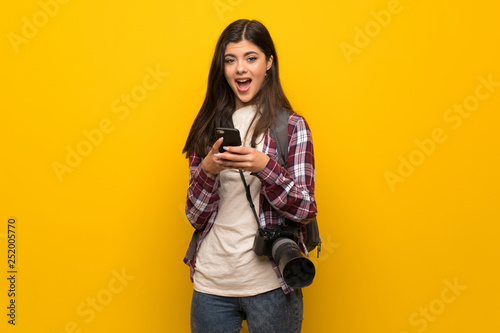 Photographer teenager girl over yellow wall surprised and sending a message