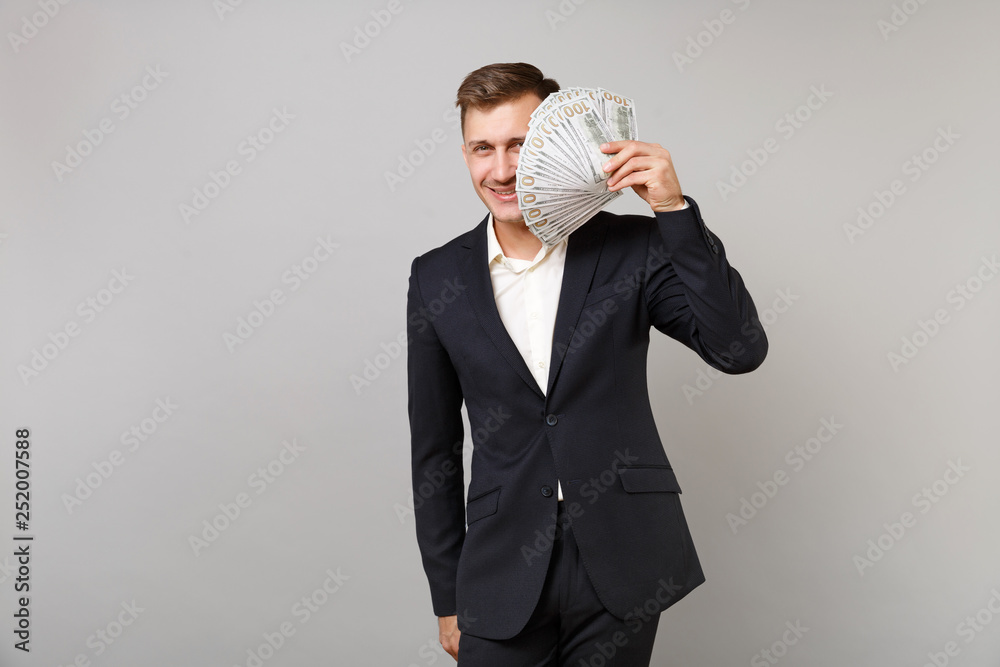 Smiling young business man in classic suit covering face with lots bunch of dollars banknotes, cash money isolated on grey background. Achievement career wealth business concept. Mock up copy space. Smiling young business man in classic suit covering face with lots bunch of dollars banknotes, cash money isolated on grey background. Achievement career wealth business concept. Mock up copy space.