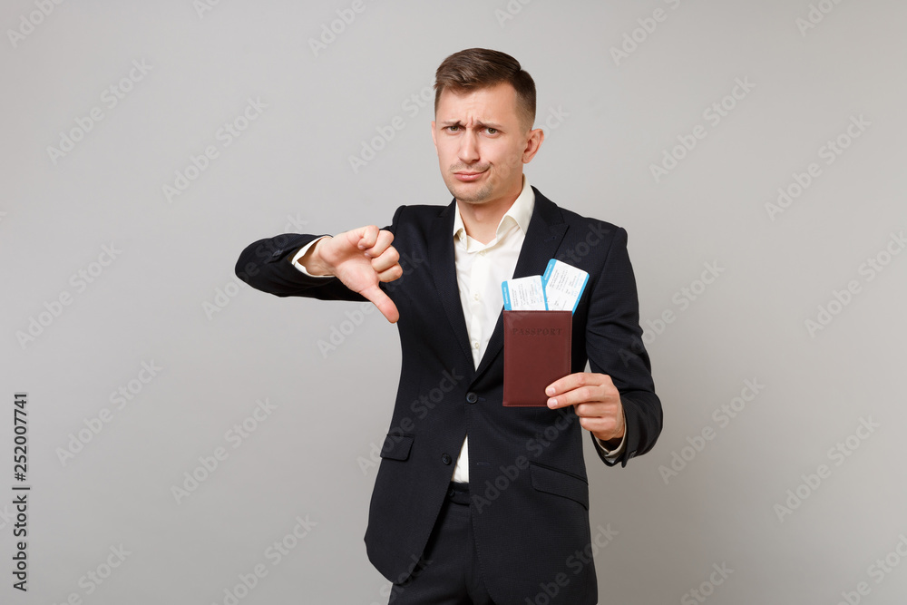 Dissatisfied young business man in classic suit showing thumb down, holding passport, boarding pass ticket isolated on grey background. Achievement career wealth business concept. Mock up copy space. Dissatisfied young business man in classic suit showing thumb down, holding passport, boarding pass ticket isolated on grey background. Achievement career wealth business concept. Mock up copy space.