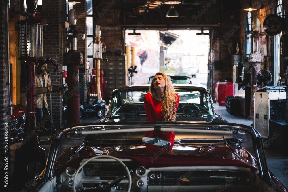 Sexy blond woman in red dress near retro car in old garage. Young sensual model posing for retro car show