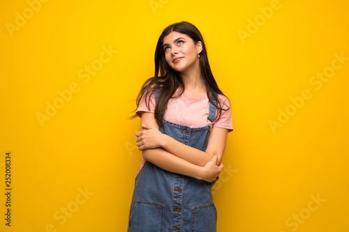 Teenager girl over yellow wall looking up while smiling