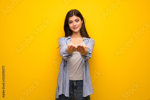 Teenager girl over yellow wall holding copyspace imaginary on the palm to insert an ad © luismolinero