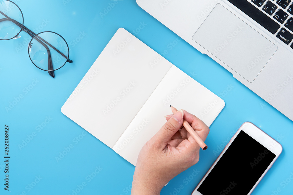 A student write on open white book or accounting isolated on a minimal clean blue workplace at home with smartphone and accessories, copy space, flat lay, top view, mock up