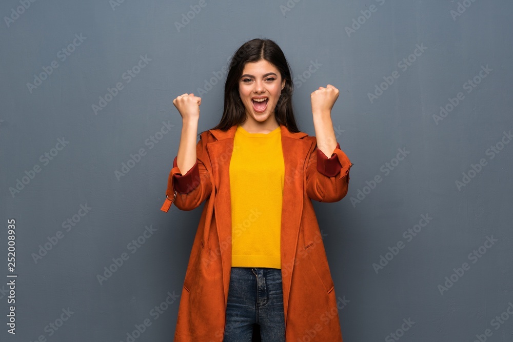 Teenager girl with coat over grey wall celebrating a victory in winner position Teenager girl with coat over grey wall celebrating a victory in winner position
