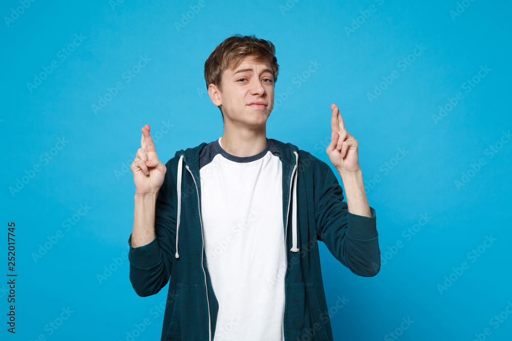 Confused young man in casual clothes waiting for special moment, keeping fingers crossed, making wish isolated on blue wall background. People sincere emotions, lifestyle concept. Mock up copy space. Confused young man in casual clothes waiting for special moment, keeping fingers crossed, making wish isolated on blue wall background. People sincere emotions, lifestyle concept. Mock up copy space.