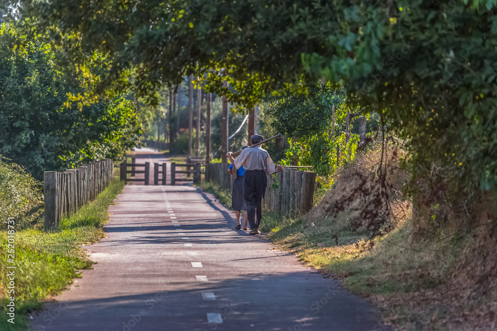 View of eco pedestrian / cycle lane, with couple of senior farmers, walking and carrying agricultural tools