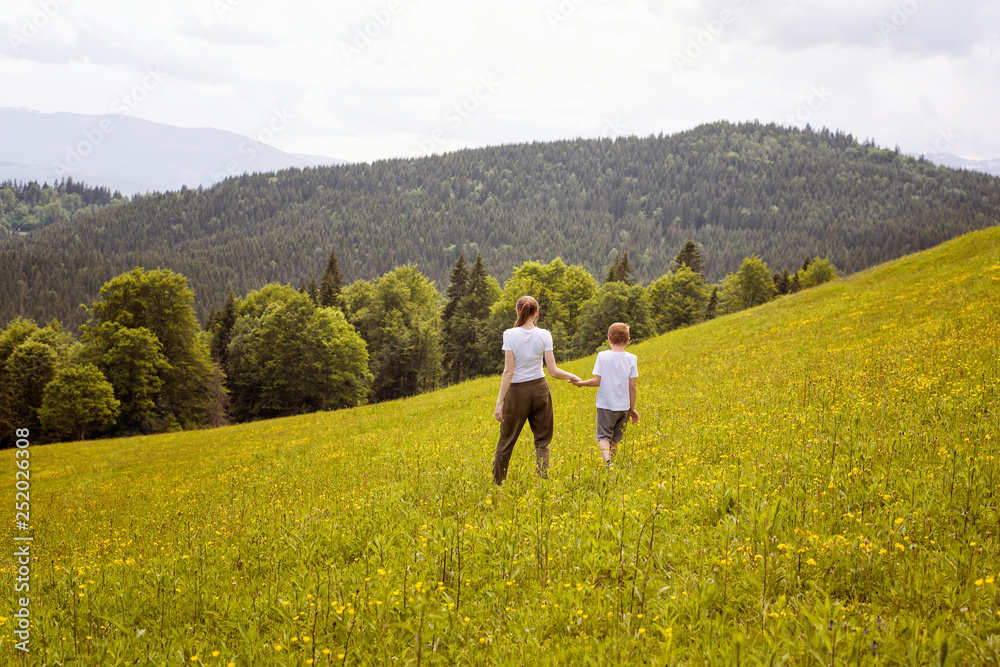 Mother little son go hand in hand on a green field against the background of the forest, mountains and sky with clouds.
