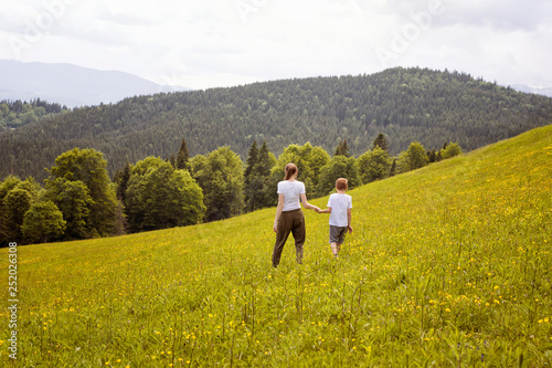 Mother little son go hand in hand on a green field against the background of the forest  mountains and sky with clouds.