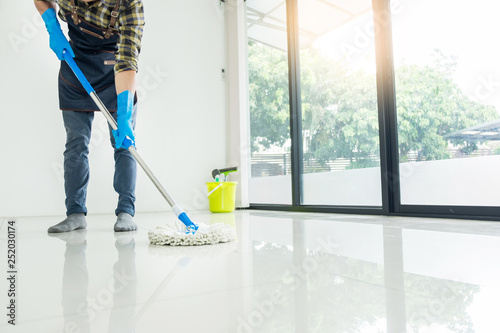 Young housekeeper cleaning floor mobbing holding mop and plastic bucket with brushes, gloves and detergents in the leaving room house floor helping his wife.