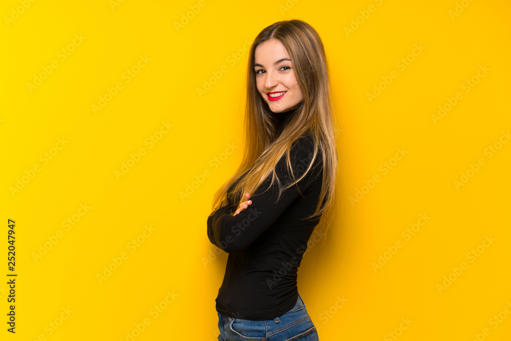 Young pretty woman over yellow background keeping the arms crossed in lateral position while smiling