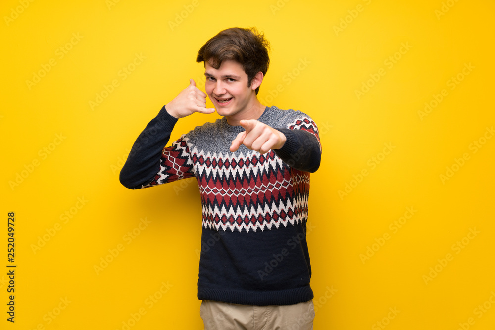 Teenager man over yellow wall making phone gesture and pointing front Teenager man over yellow wall making phone gesture and pointing front