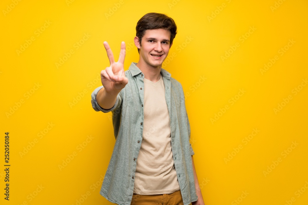 Teenager man over yellow wall smiling and showing victory sign Teenager man over yellow wall smiling and showing victory sign