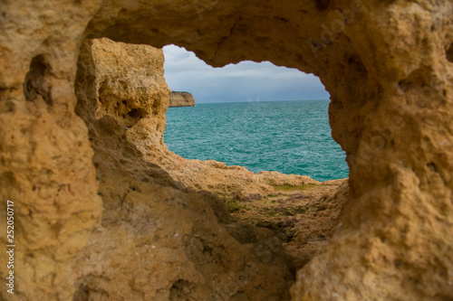 The hole is a cave in the yellow rock, through which you can see the blue water of the Atlantic Ocean and the precipice of the opposite shore in the Algarve, Portugal. © Alona