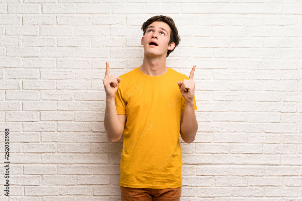 Teenager man over white brick wall pointing up and surprised Teenager man over white brick wall pointing up and surprised