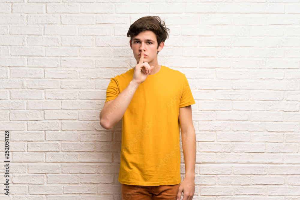 Teenager man over white brick wall showing a sign of silence gesture putting finger in mouth