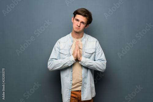 Teenager man with jean jacket over grey wall keeps palm together. Person asks for something
