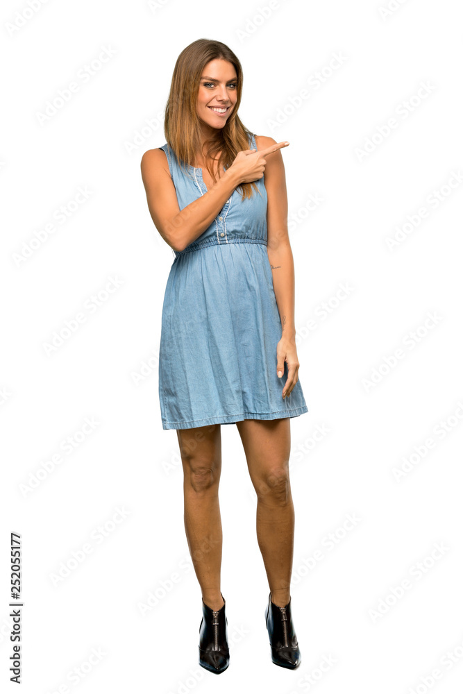 A full-length shot of a Blonde woman with jean dress pointing to the side to present a product over isolated white background A full-length shot of a Blonde woman with jean dress pointing to the side to present a product over isolated white background